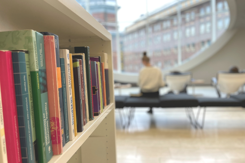 A bookshelf in the foreground, in the background a student studying in front of a window.