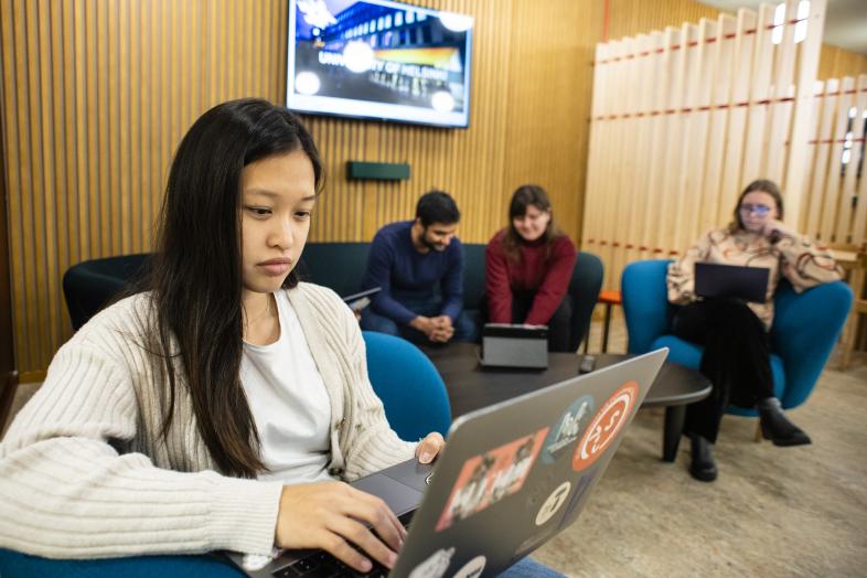 opiskelijoita istuskelemassa aulatilassa, kannettavat tietokoneet sylissä.