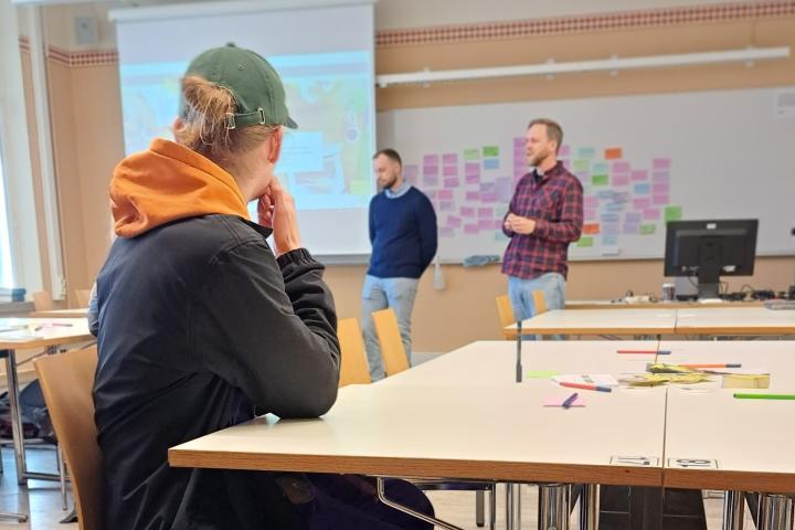 A student looking towards a whiteboard, two teachers in the background.